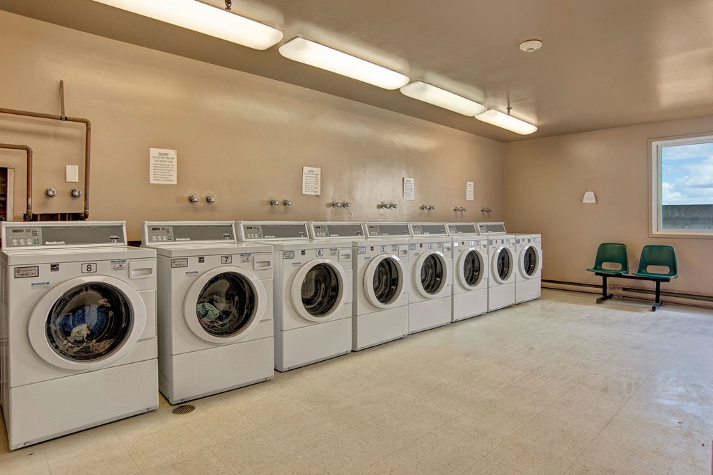 A bright, clean laundromat here at Centre Pointe Plaza Apartment Homes with nine white front-loading washing machines lined up. Two green chairs sit by a window, creating a tidy, orderly atmosphere.