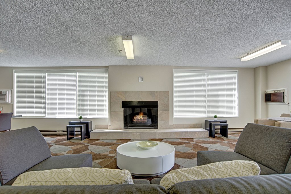 Modern resident lounge here at Centre Pointe Plaza Apartment Homes with a cozy fireplace between large windows with blinds. Gray sofas face a circular white coffee table, creating a calm atmosphere.