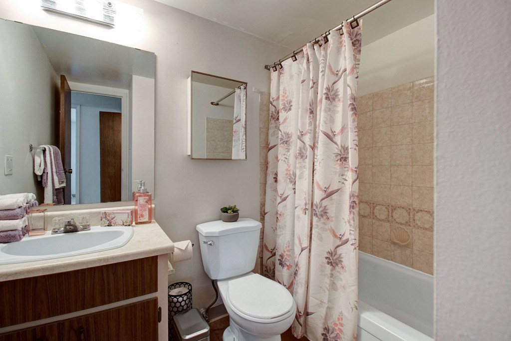 Compact bathroom here at Centre Pointe Plaza Apartment Homes with beige tiles, floral shower curtain, and wood vanity with sink. Soft lighting, towels, and soap create a cozy, tidy atmosphere.