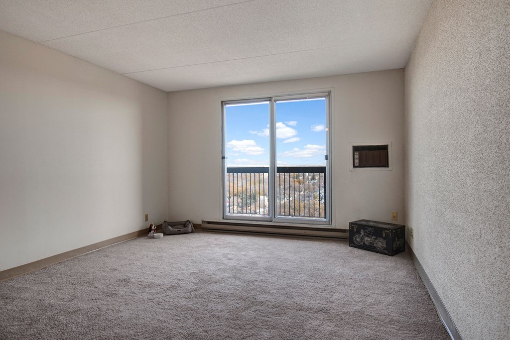 Empty bedroom here at Centre Pointe Plaza Apartment Homes with beige carpet and white walls, featuring a large window overlooking a scenic view. A small air conditioner is built into the wall, and a decorative box sits on the floor. The atmosphere feels calm and spacious.