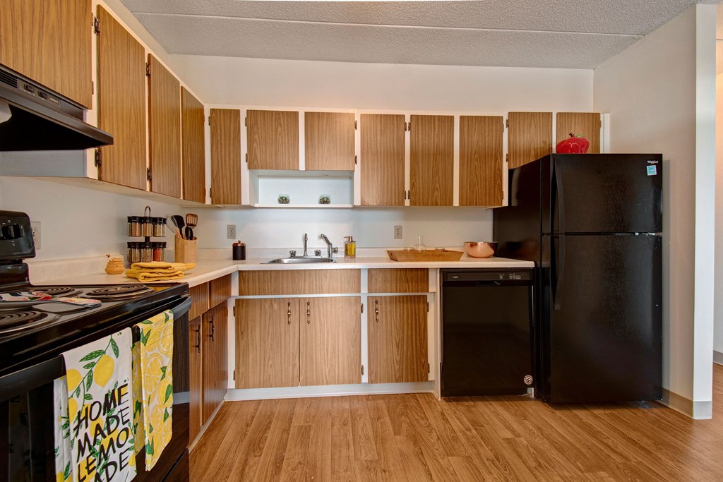 A cozy kitchen here at Centre Pointe Plaza Apartment Homes with wood cabinets, black appliances, and a light countertop. A vibrant lemon-patterned towel hangs on the oven, adding a cheerful touch.