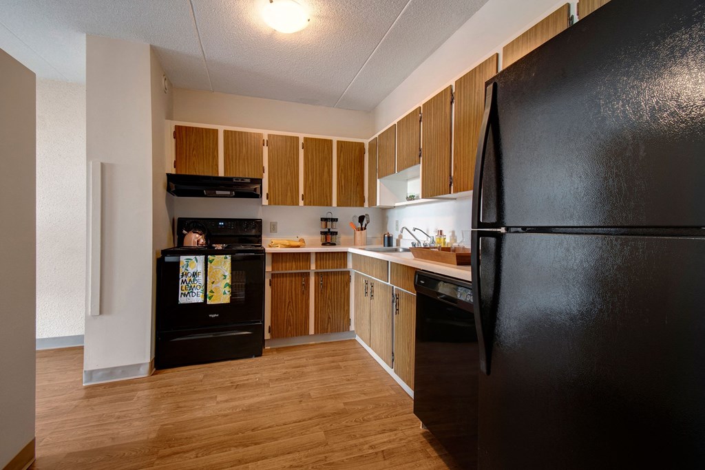 Compact kitchen here at Centre Pointe Plaza Apartment Homes with wood cabinets, black appliances, and a wooden floor. Counters hold utensils and containers; cozy and organized ambiance.