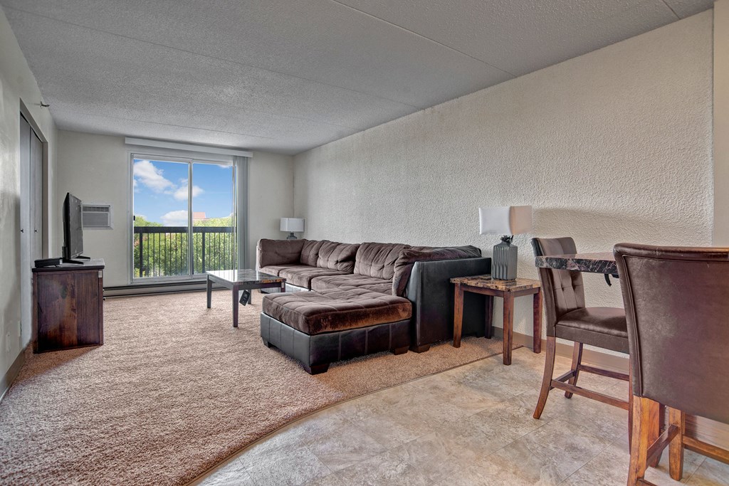 Spacious living room here at Centre Pointe Plaza Apartment Homes with a large brown sectional sofa, a coffee table, and a TV on a stand. Sliding glass door reveals a view of trees outside.