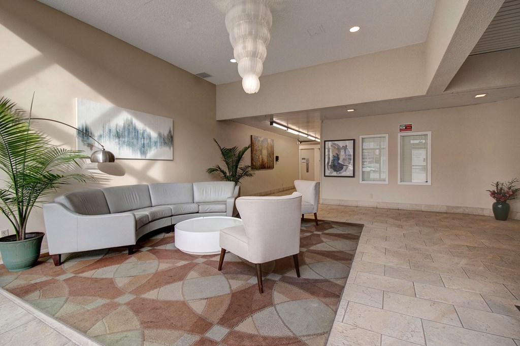 A modern lounge here at Centre Pointe Plaza Apartment Homes with a curved gray sofa and two white armchairs on a patterned rug. Potted plants and abstract art create a calm, welcoming atmosphere.