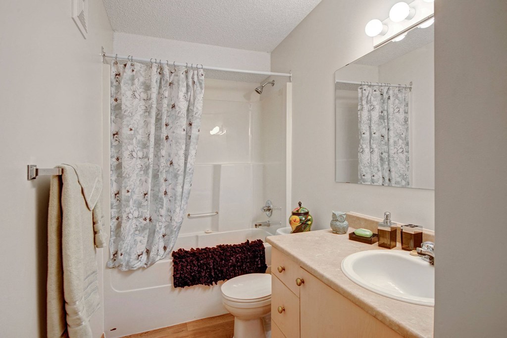 Bathroom here at Edgewood Estates Apartment Homes with a floral shower curtain, beige walls, and a wooden vanity with a sink. A mirror reflects light from above, creating a cozy ambiance.