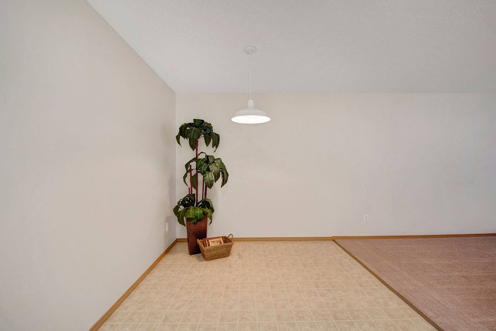 Simple room here at Edgewood Estates Apartment Homes with light beige walls, a potted plant in the corner, and a woven basket. A white ceiling light hangs above tiled and carpeted flooring.