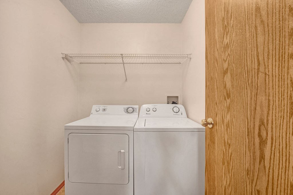 Laundry room here at Edgewood Estates Apartment Homes with a white washer and dryer side by side under a wire shelf on a beige wall. A wooden door is partially open on the right.