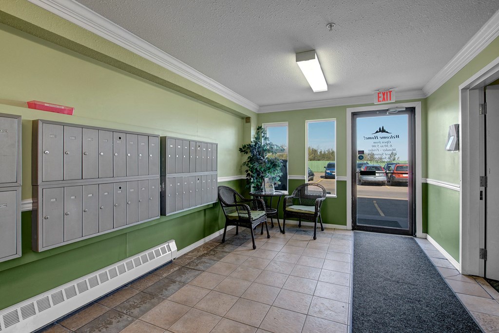 Edgewood Estates Apartment Homes lobby with pale green walls, a wall of mailboxes, and wicker chairs around a small table featuring a plant. A glass door leads outside.