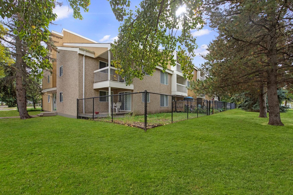 Gateway Gardens Apartment Homes complex with beige exterior and balconies, surrounded by a black fence and lush green lawn. Large trees provide shade, creating a serene atmosphere.