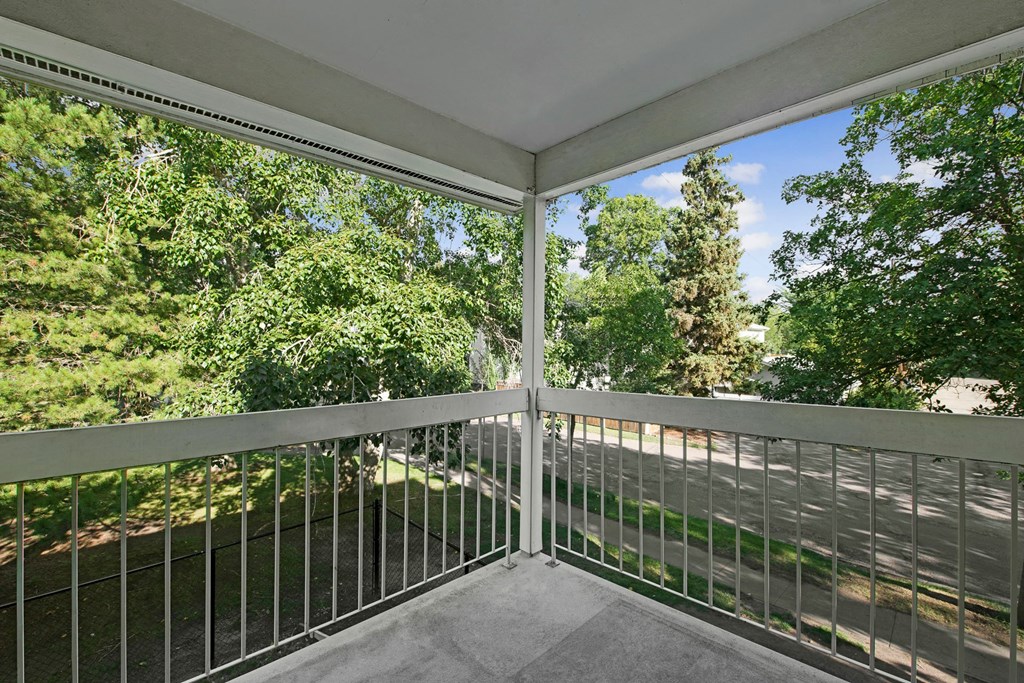Covered balcony here at Gateway Gardens Apartment Homes with metal railing overlooks lush green trees and a quiet street. The scene feels serene and inviting under a clear blue sky.
