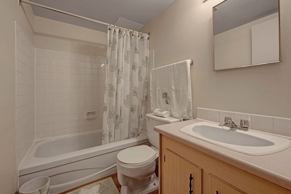 Small bathroom here at Gateway Gardens Apartment Homes with a bathtub and shower curtain on the left, toilet in the center, and wooden cabinet with sink and mirror on the right. Bright and clean atmosphere.