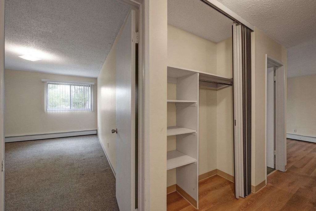 Empty room here at Gateway Gardens Apartment Homes with beige walls and carpet, illuminated by a square ceiling light. Open closet with shelves on the right. Window with vertical blinds reveals greenery outside.
