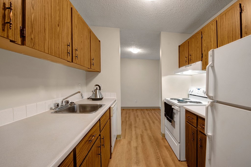 Narrow kitchen here at Gateway Gardens Apartment Homes with wooden cabinets, white countertops, stainless steel sink, and kettle. White appliances include a fridge and stove. Cozy and simple.