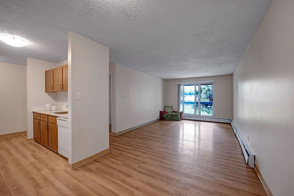 Empty apartment here at Gateway Gardens Apartment Homes with wood flooring, an open kitchen with wooden cabinets, and bright natural light from large windows. Serenity, simplicity, and modernity.