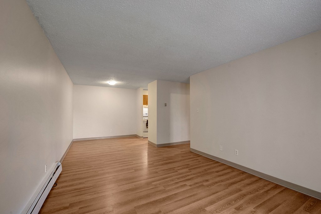 Empty living room here at Gateway Gardens Apartment Homes with light brown wood flooring and beige walls, illuminated by a ceiling light. An open doorway leads to a small kitchenette. Minimalistic and clean.