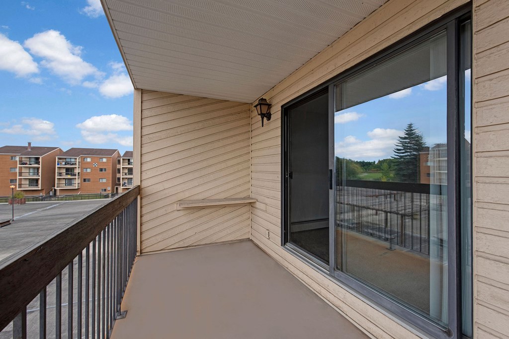 Apartment balcony here at Gateway Square Apartment Homes with beige siding and sliding glass doors, overlooking nearby residential buildings and greenery. Clear blue sky with fluffy clouds.