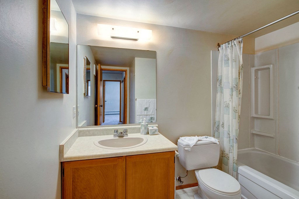 Simple bathroom here at Gateway Square Apartment Homes with a wood cabinet sink, large mirror, white toilet, and a tub with a shower curtain. Neutral tones create a clean and calm vibe.