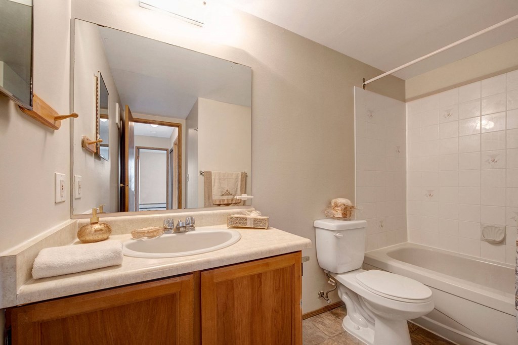 Simple bathroom here at Gateway Square Apartment Homes with wooden vanity, oval mirror, and beige countertop. White toilet and bathtub. Soft lighting creates a clean, inviting atmosphere.