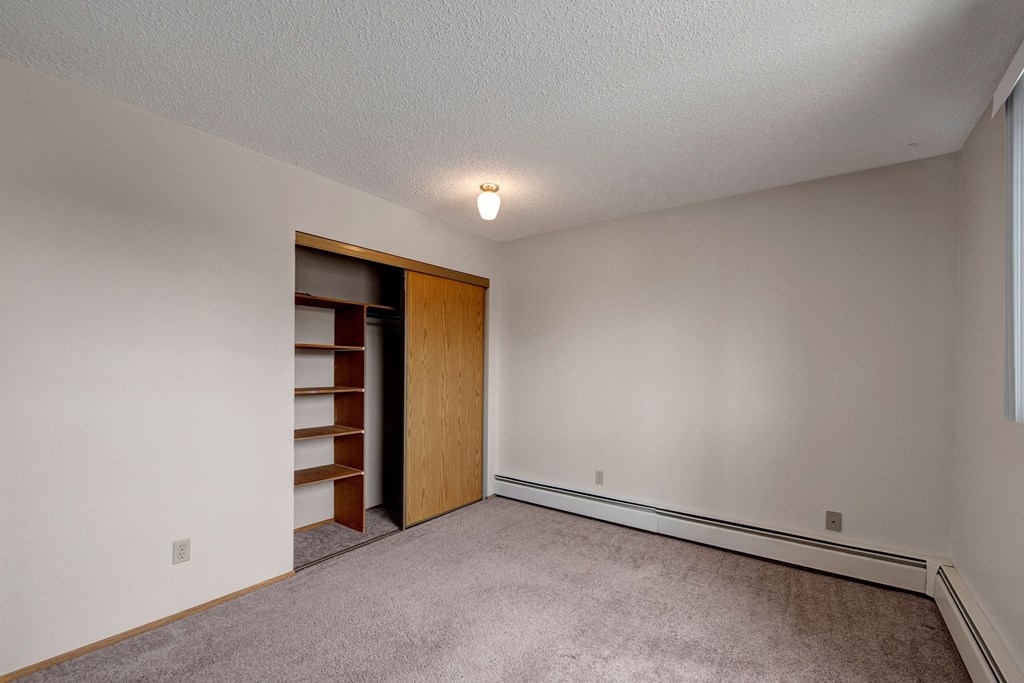 Empty bedroom here at Gateway Square Apartment Homes with beige walls, a small ceiling light, and carpeted floor. Open closet with shelves on the left, creating a clean, neutral atmosphere.