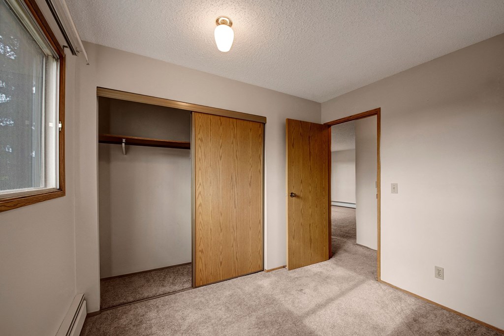 Small, empty room here at Gateway Square Apartment Homes with beige walls and carpet, a light fixture, a wooden closet with sliding doors, and a window letting in natural light.