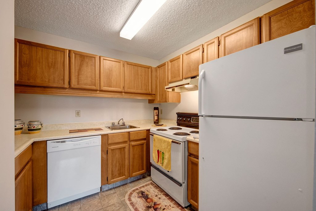 Cozy kitchen here at Gateway Square Apartment Homes with wooden cabinets, white appliances, and beige countertops. Features include a fridge, stove, dishwasher, and decorative rug. Simple and neat.