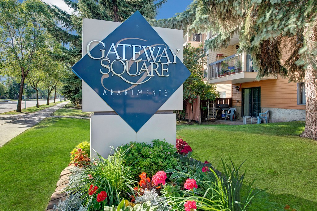 Sign reading "Gateway Square Apartments" in front of a well-maintained lawn with colorful flowers, trees, and a tan apartment building in the background.
