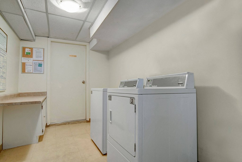 Laundry room here at Kingsmere Apartment Homes with two white industrial washers against a plain wall. A door and bulletin board with notices are on the left. The space is bright and clean.