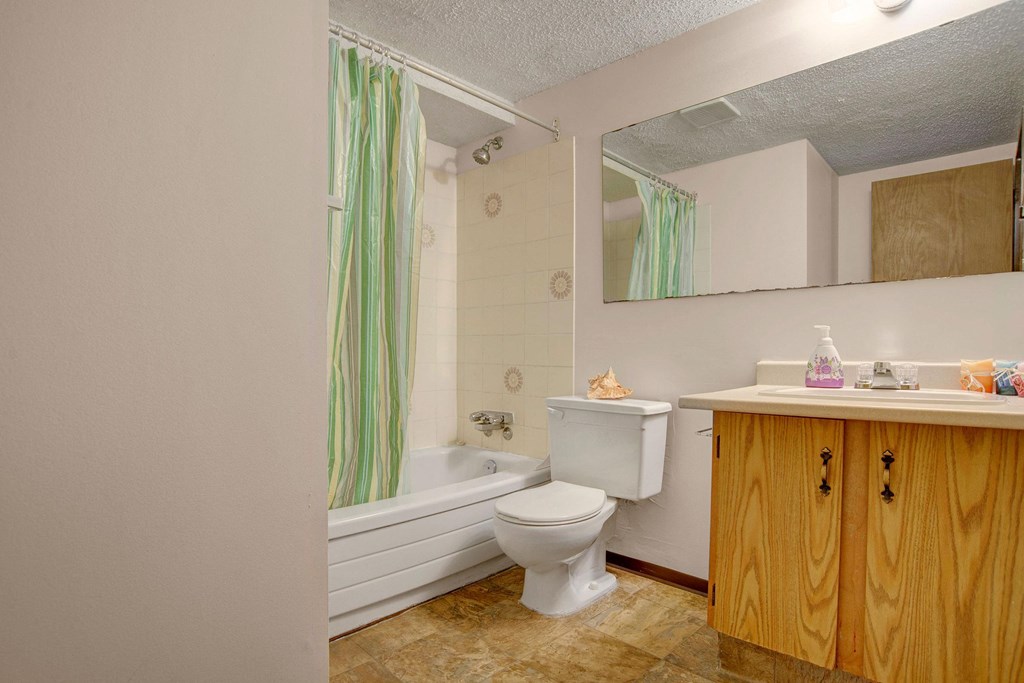 Bathroom here at Kingsmere Apartment Homes with a bathtub and green striped shower curtain, wooden vanity with a large mirror, and a toilet. The atmosphere is clean and simple.
