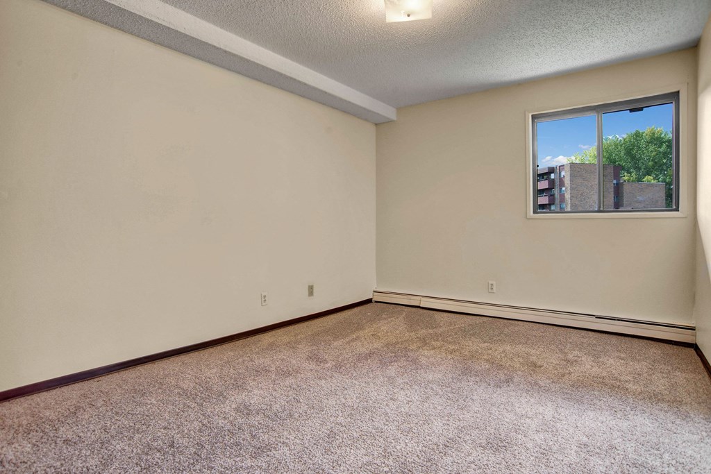 Empty bedroom here at Kingsmere Apartment Homes with beige walls, carpeted floor, and a single window showing a clear blue sky and green trees. The atmosphere is quiet and neutral.