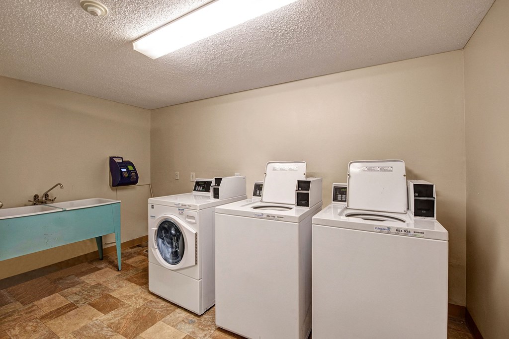 Laundry room here at Lakewood Heights Apartment Homes with two white top-load washing machines and a front-load dryer. A utility sink with a faucet is on the left. Beige walls and tiled floor.