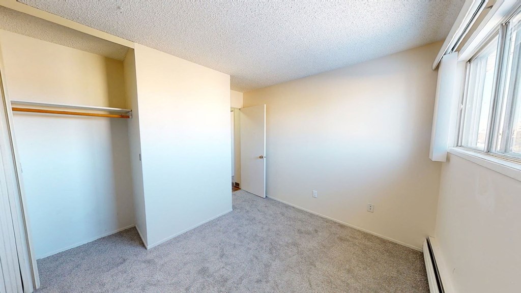 Empty room here at Lakewood Heights Apartment Homes with beige walls, gray carpet, and a large window on the right. An open closet with a wooden rod and a partially open white door is visible. Bright, airy feel.
