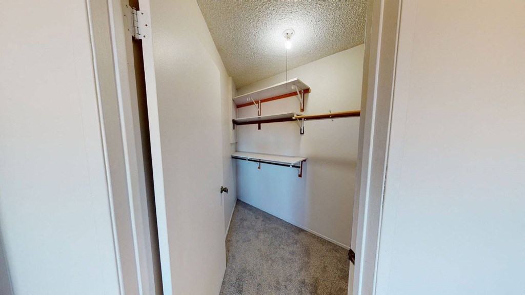 A small walk-in closet here at Lakewood Heights Apartment Homes with beige carpet, white walls, and a single ceiling light. Two wooden rods and white shelves for storage are visible.