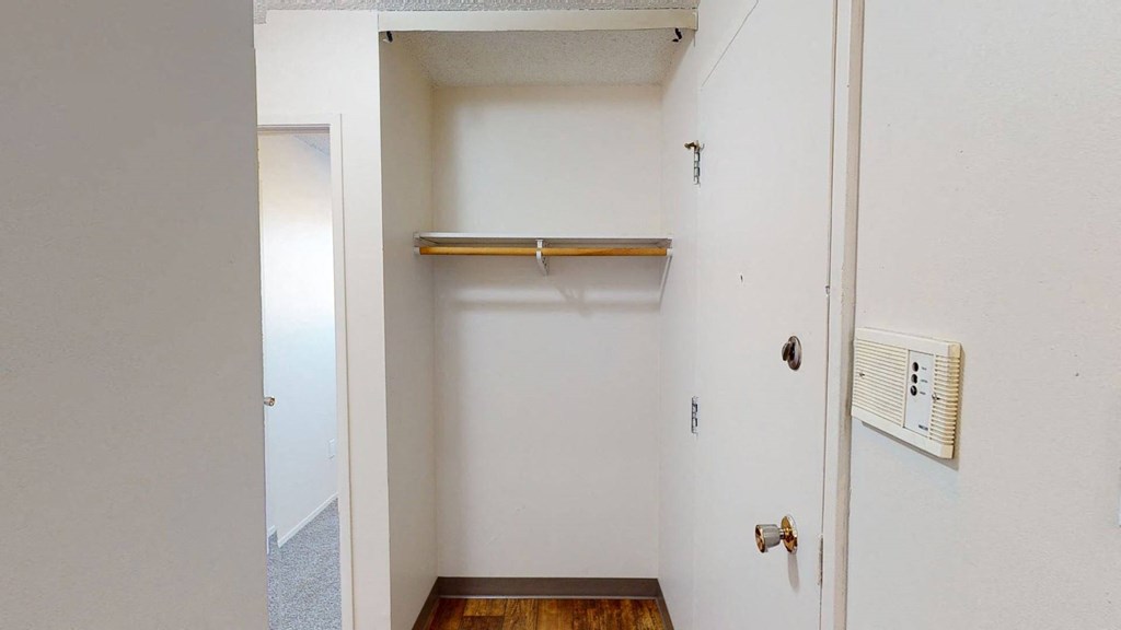 Empty white closet here at Lakewood Heights Apartment Homes with a wooden rod and shelf, adjacent to a door with a round knob. A light switch and intercom are mounted on the wall.