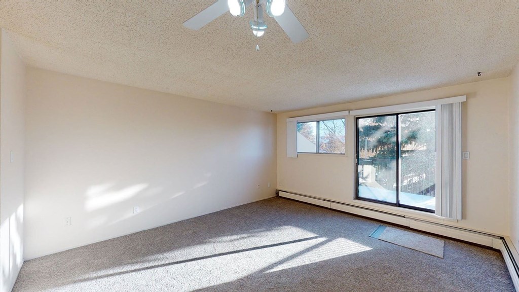 Empty room here at Lakewood Heights Apartment Homes with beige walls, carpeted floor, and ceiling fan. Large sliding glass doors and a window let in sunlight, casting shadows across the room.