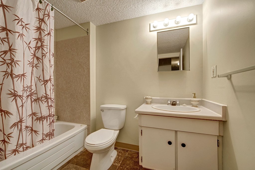 Compact bathroom here at Lakewood Heights Apartment Homes with beige walls, featuring a bamboo-patterned shower curtain, white toilet, and sink with a mirror above. Soft, warm lighting.