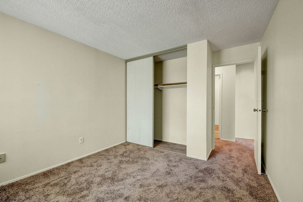 Empty bedroom here at Lakewood Heights Apartment Homes with beige walls and carpet, featuring an open closet with sliding doors. A partially open door reveals a hallway, creating a calm, neutral tone.