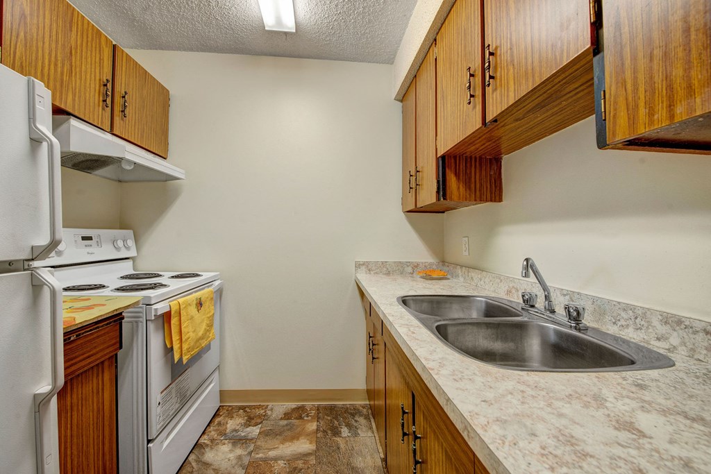 Compact kitchen here at Lakewood Heights Apartment Homes with white appliances and brown wooden cabinets. Marble countertops with a double sink and a yellow towel on the stove, creating a cozy feel.