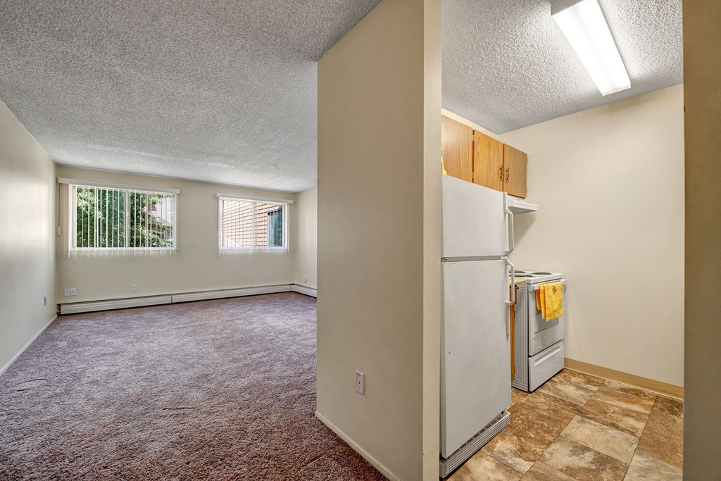 Spacious empty room here at Lakewood Heights Apartment Homes with beige carpet and two large windows with vertical blinds. Adjacent kitchen with white appliances and yellow towel. Bright and minimal.