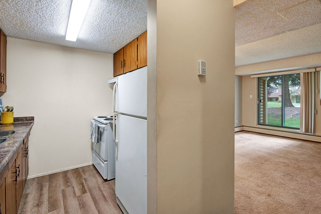 Compact kitchen here at Lakewood Heights Apartment Homes with wood cabinets and a white stove, adjacent to a bright living room with beige carpet and large window showing a grassy view. Cozy atmosphere.