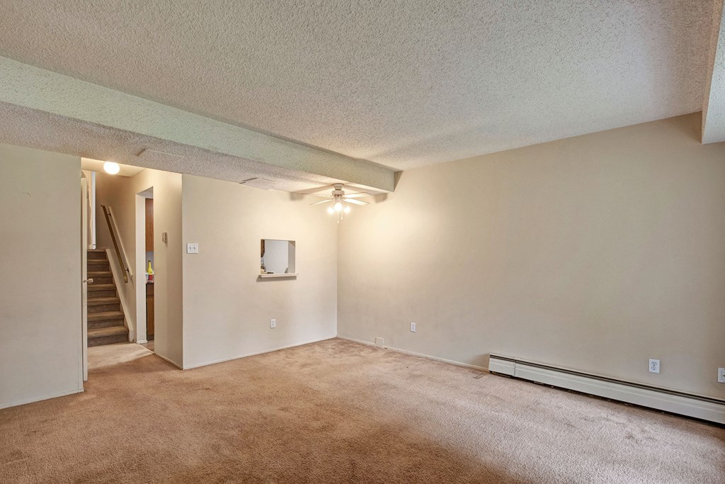 Empty living room here at Lakewood Heights Apartment Homes with beige walls and carpet, ceiling fan, and a small window opening to a kitchen area. Stairs are visible in the background.