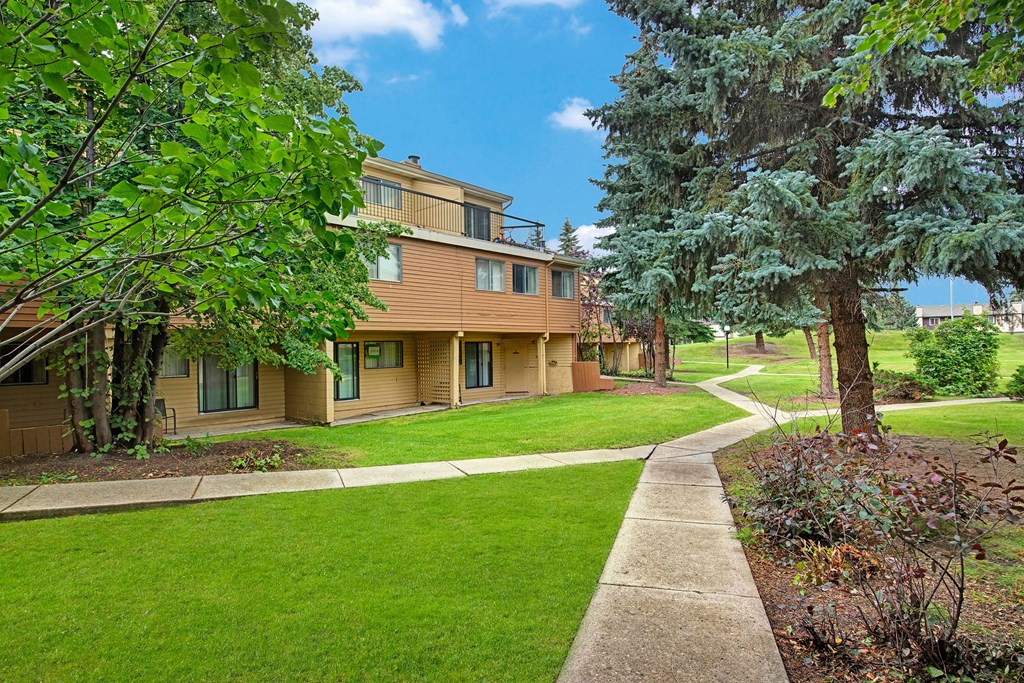 Spacious green backyard here at Lakewood Heights Apartment Homes with a beige two-story house, surrounded by lush trees and manicured lawns. A winding concrete path leads through the serene landscape.