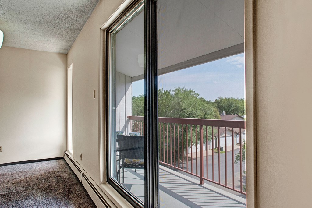 A bright room here at Madison Park Apartment Homes with a large sliding glass door leads to a balcony, revealing a suburban neighborhood and lush green trees under a blue sky.