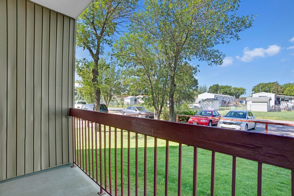 View from a balcony here at Madison Park Apartment Homes, with vertical siding and wooden railing, overlooking parked cars and trees. Mobile homes and blue sky complete the peaceful suburban scene.