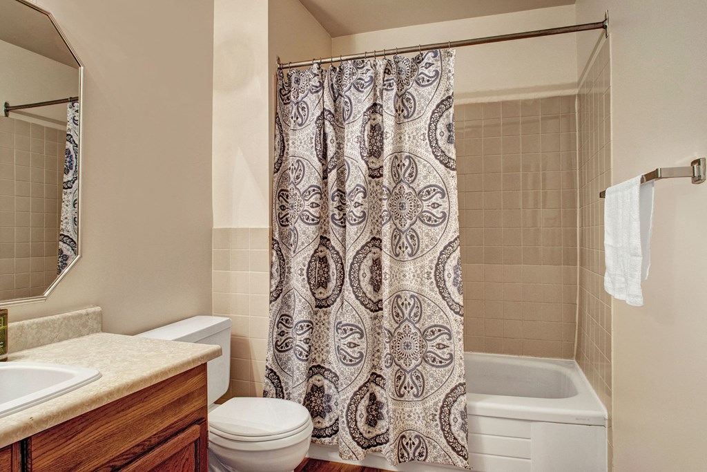 Bathroom here at Madison Park Apartment Homes featuring a tiled shower with a patterned curtain, a white toilet, a sink with a wooden cabinet, and a towel on a rack. Clean and tidy.