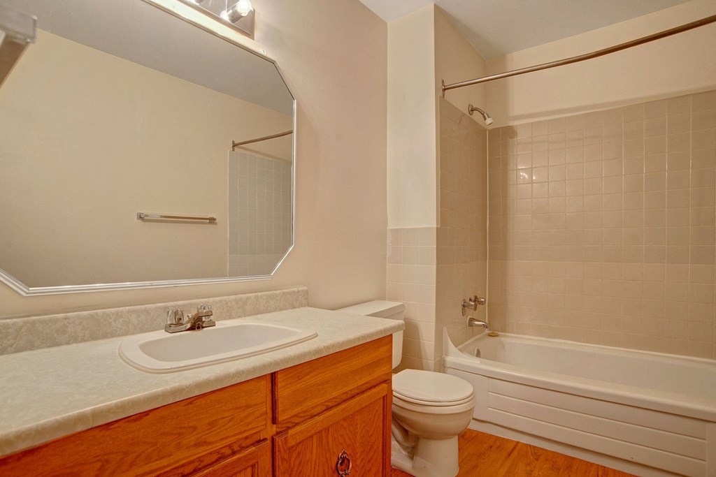 Simple bathroom here at Madison Park Apartment Homes with a large mirror above a white countertop and wooden cabinets. Beige tiled bathtub, shower, and toilet, conveying a clean, neutral tone.