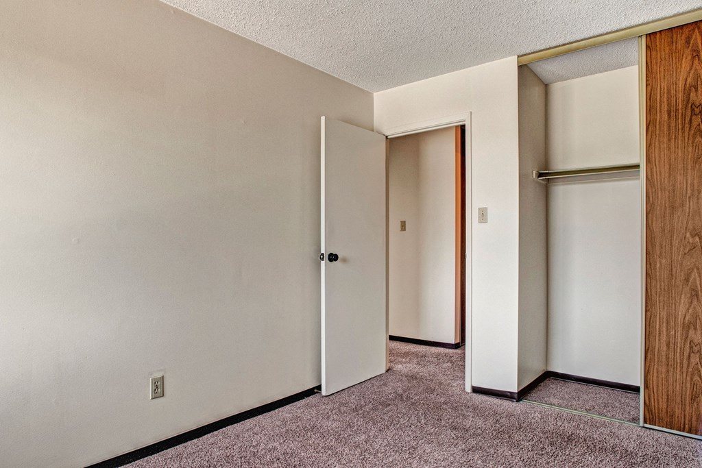 Empty room here at Madison Park Apartment Homes with beige walls, brown carpet, and an open white door leading to a hall. A closet with sliding wooden doors is on the right.