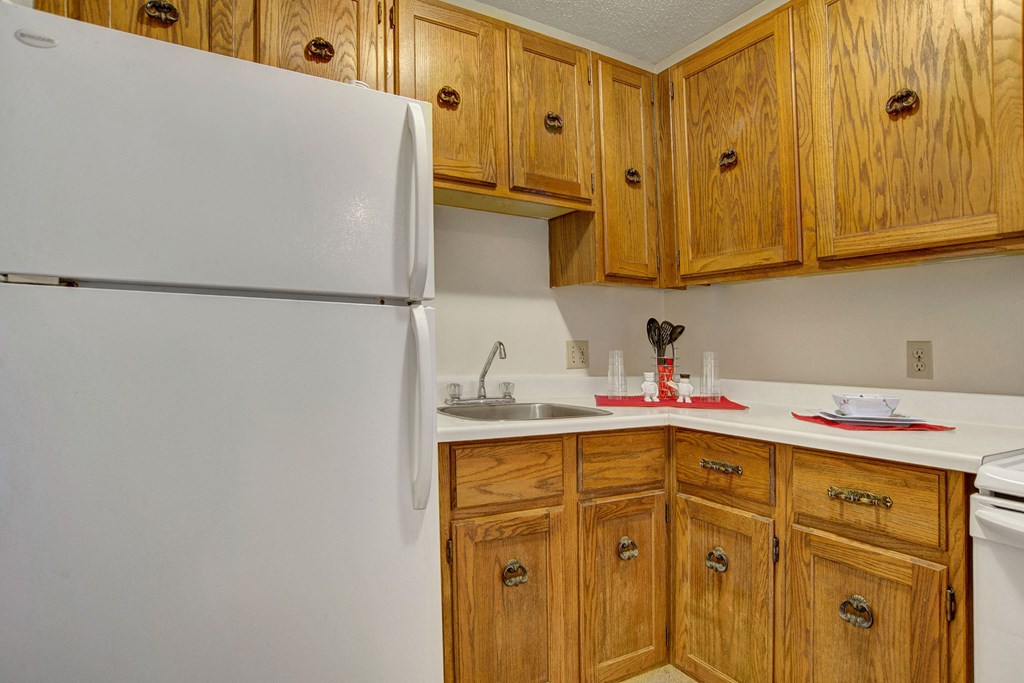 A cozy kitchen here at Madison Park Apartment Homes features wooden cabinets, a white refrigerator, and a sink. Countertops hold utensils, cups, and plates, creating a warm, inviting atmosphere.