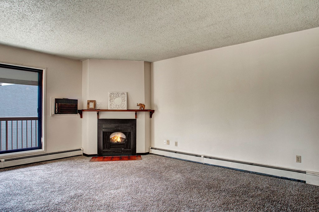 Spacious living room here at Madison Park Apartment Homes with beige walls and carpet, featuring a cozy fireplace with a wooden mantel. Large window provides natural light. Minimalist decor.