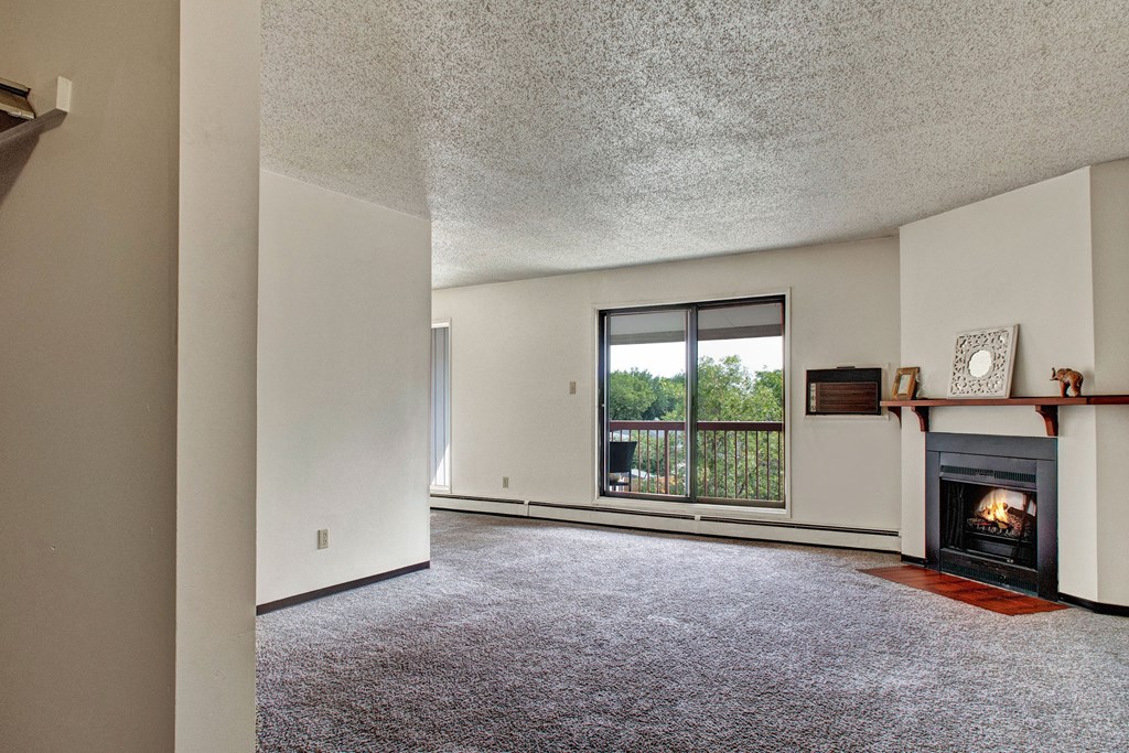 Spacious, carpeted living room here at Madison Park Apartment Homes with white walls, a cozy lit fireplace, and a large window revealing green trees outside, creating a serene ambiance.
