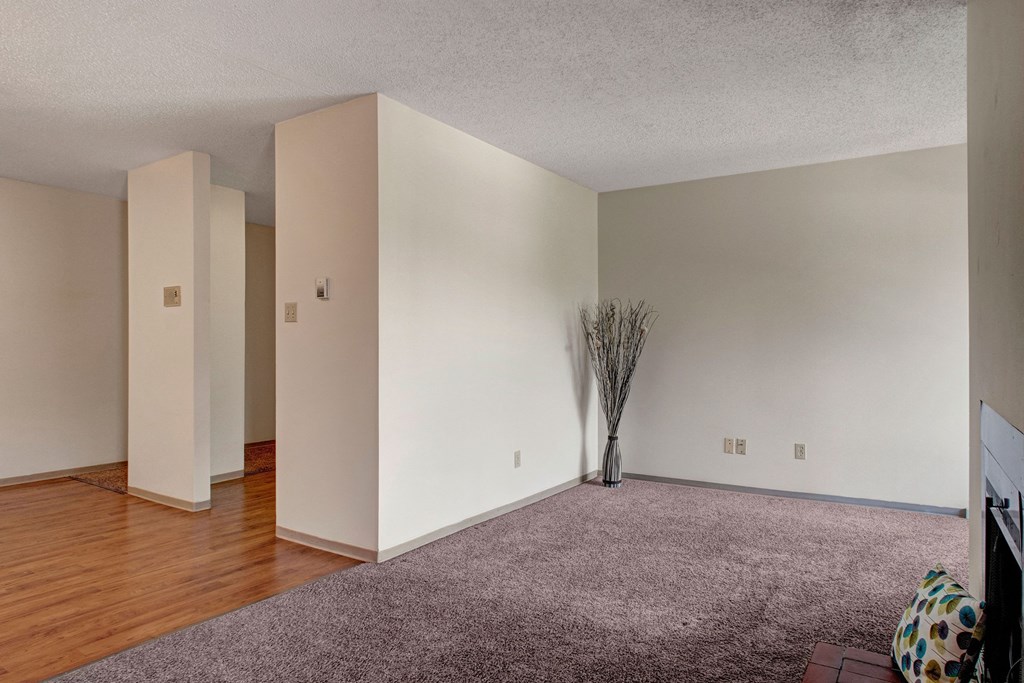 Spacious empty room here at Madison Park Apartment Homes with beige walls, featuring a blend of carpet and hardwood flooring. A vase with tall branches adds a touch of decor.